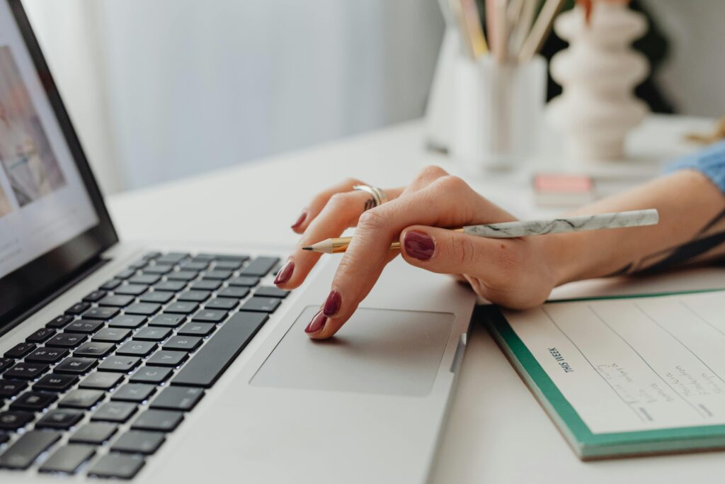 A close-up shot of a woman's hand on a laptop touchpad, holding a pencil, beside a notebook.